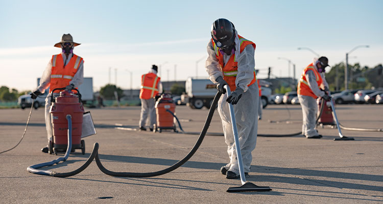 workers in protective gear using large vacuum cleaners to clean an asphalt surface