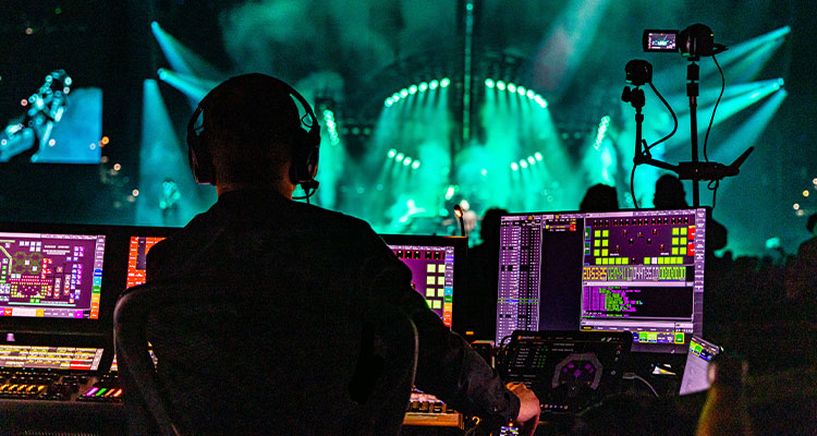 a live event production setup. A technician is seen from behind, wearing headphones and operating a complex control desk with multiple computer screens