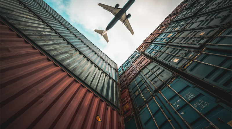 A commercial airplane flying above stacked shipping containers, symbolising global cargo transport and supply chain logistics